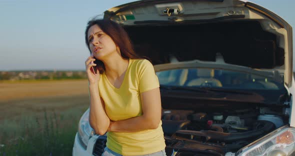 Upset Young Woman Driver Talking Angrily on Cell Phone with Assistance Service Near a Broken Car