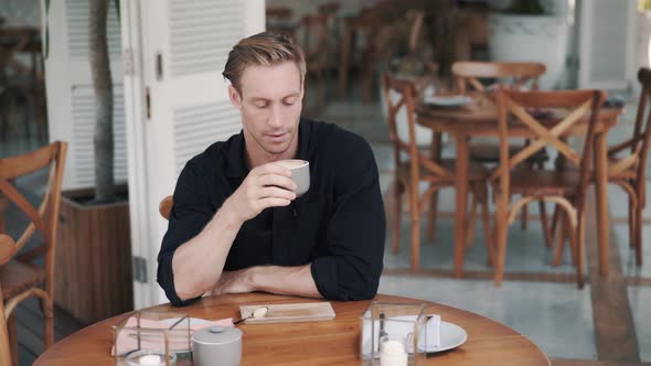 Handsome Man Drinks Coffee in Cafe and Puts Sugar in Beverage, Stirs with Spoon