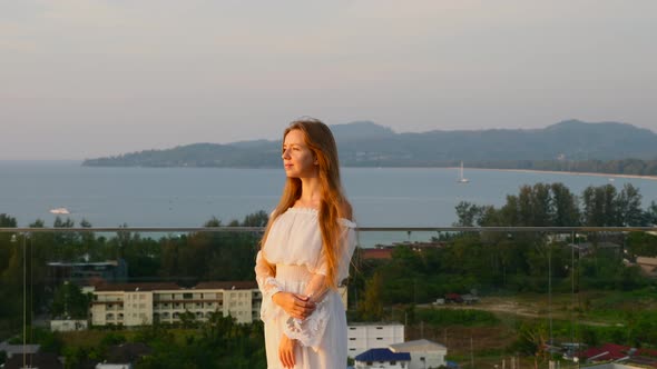 Smiling Woman in White Dress Standing on Rooftop with Sea Sunset and Mountain