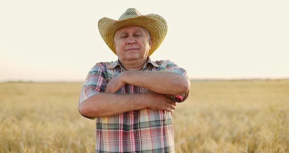 Happy Farmer in Straw Hat Standing in Wheat Field at Sunset Crossing His Arms on Breast and Smiling