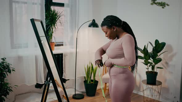 Positive African American Woman Measuring Her Waist with Tape Against Mirror and Enjoying Result of