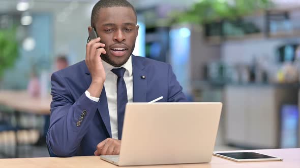 African Businessman with Laptop Celebrating on Smartphone in Office