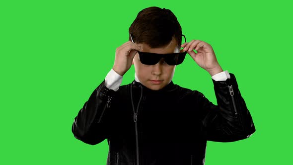 Cool Young Boy in a Leather Jacket Putting on Black Glasses with Arms on His Hips on a Green Screen