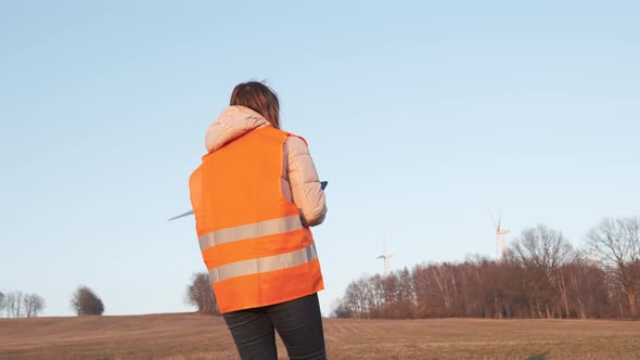 Female Engineer in Orange Vesta Goes to Wind Turbines with a Tablet to Checks Their Operation