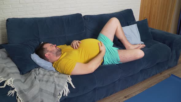 Plump Man with Beard in Yellow Tshirt is Lying on Bed at Home and Stroking Big Sick Stomach