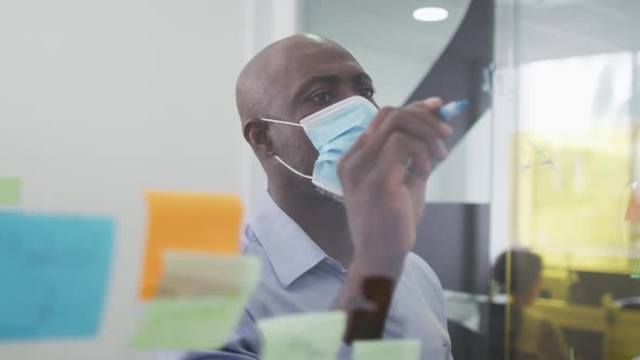 African american businessman in face mask brainstorming, writing on board with memo notes in office