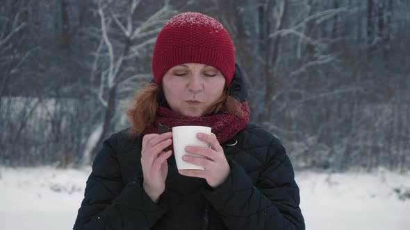 Woman in a red warm hat is drinking hot tea or coffee from a cup in a snowy forest