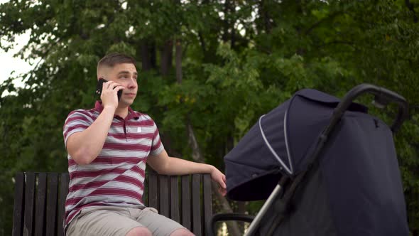 A Young Father is Sitting on a Bench and Talking on the Phone with a Stroller