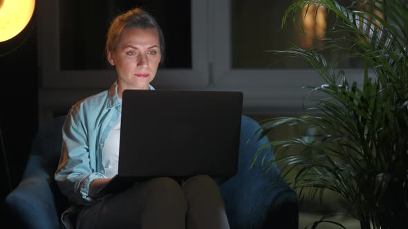 Woman is Sitting in the Armchair and Working on a Laptop at Night