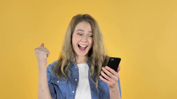 Young Pretty Girl Excited for Success While Using Smartphone on Yellow Background