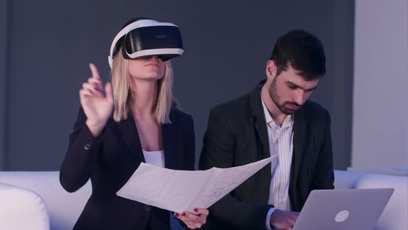 Blonde Woman in VR Headset Holding Construction Plans While Her Male Partner Typing on the Laptop