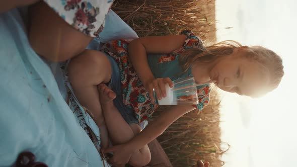 A Cute Preschool Girl Sits Crosslegged and Drinking Milk Through a Straw at a Family Picnic in a