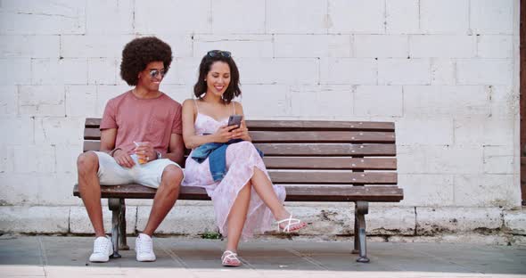Two Smiling People Sitting on Bench Resting Using Smartphone. Tourists Resting on bench