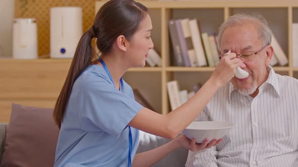Young Asian nurse feeding breakfast to senior man at couch. Old Asian man with white beard enjoy his