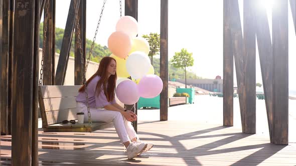 Upset Redhead Young Woman Swaying on Swing Holding Hands Large Fountain of Colorful Balloons