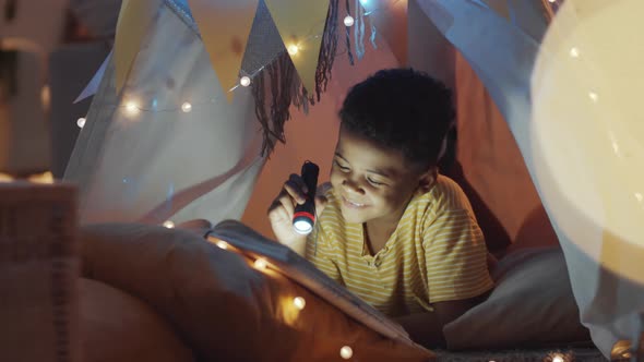 Cheerful Kid Holding Flashlight and Reading Book in Teepee at Home