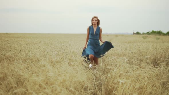 Handsome Young Woman in a Long Blue Dress Running Through Golden Wheat Field Looking to the Camera