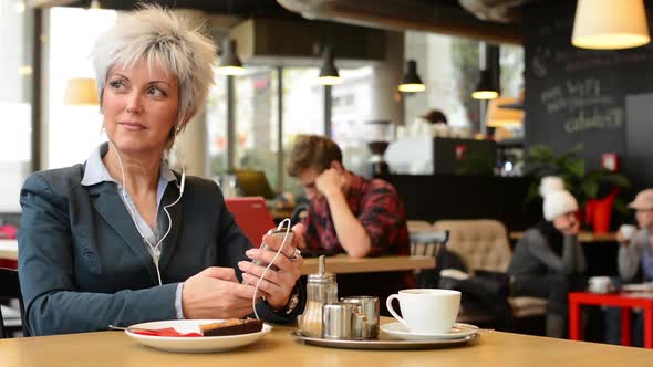 Middle Aged Woman Listens Music in Cafe (Earphone) on Smartphone