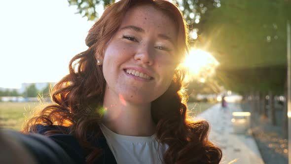 Woman with Curly Red Hair Makes Selfie Winking in City Park
