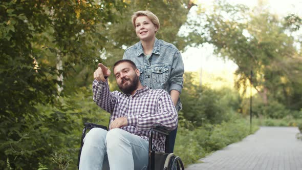 Woman Rolls Emotional Disabled Man in Wheelchair Who Points Way with His Finger Through Park Side