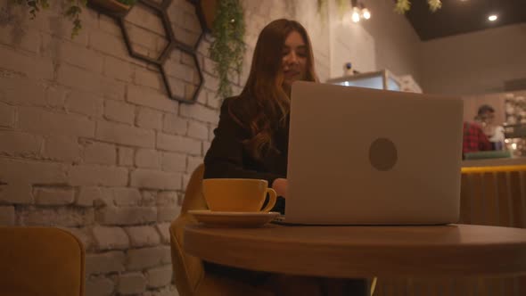 Adorable Young Girl Typing on a Laptop Keyboard in a Cafe with a Stylish Interior and Laughing at