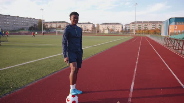 A Young Black Girl Stands on a Stadium Treadmill with Her Foot on a Soccer Ball and Points Her Index