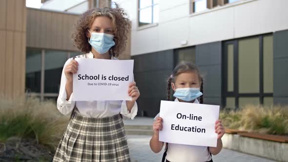 Two Schoolgirls Wearing Medical Masks with Placards Stand in Front of the School Building