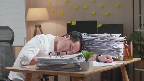 Tired Asian Man Sleeping Due To Working Hard With Documents At The Office