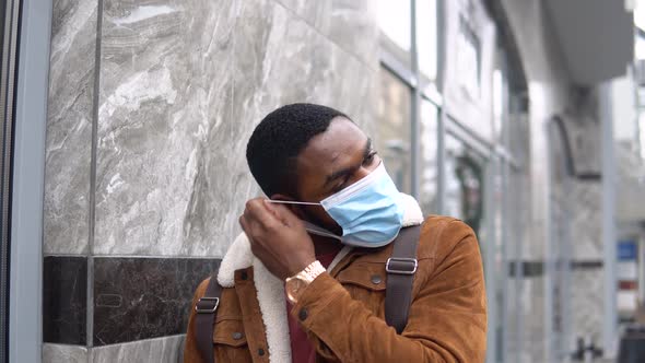 Bearded Black with Medical Mask and Shoulder Backpack Stands Outdoor Near the Office Center