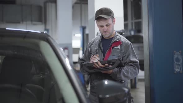 Portrait of Serious Caucasian Maintenance Engineer Checking Automobile with Digital Equipment. Adult