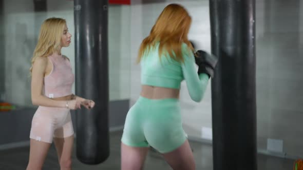 Back View of Redhead Motivated Strong Woman Boxing Punching Bag As Blond Coach Talking Endorsing