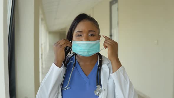 Young African American Doctor Woman in Blue Suit and White Coat Removes Disposable Sterile Face Mask