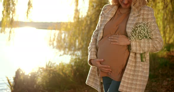 Pregnant Blonde Woman with a Bouquet on the Shore of the Pond