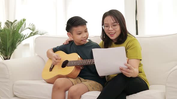 Asian mother and son sitting on the white sofa in the house son plays guitar