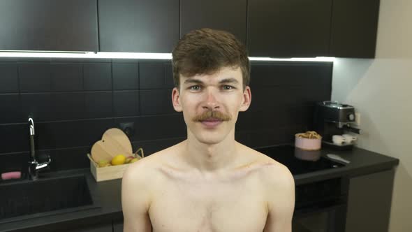 Portrait of young naked caucasian man with mustache in kitchen at home looking to camera and smiling