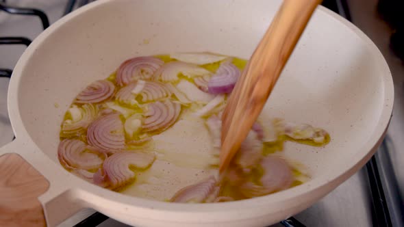 Stirring sliced raw purple onions in white frying pan with heated olive oil close up