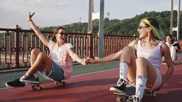 Portrait of Two Teenage Girls Riding on City Bridge Sitting on Skateboards and Holding Hands Youth