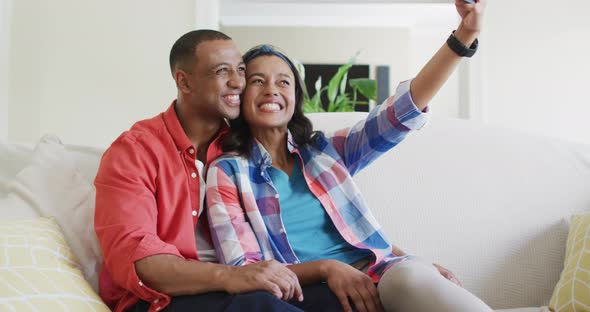 Happy biracial couple sitting on sofa, embracing, using smartphone and laughing