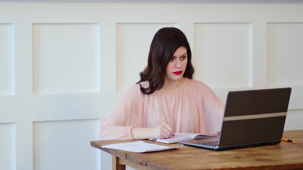 Pretty Young Lady Sitting at Desk and Typing on Laptop and Writing Tasks for the Day in a Notebook