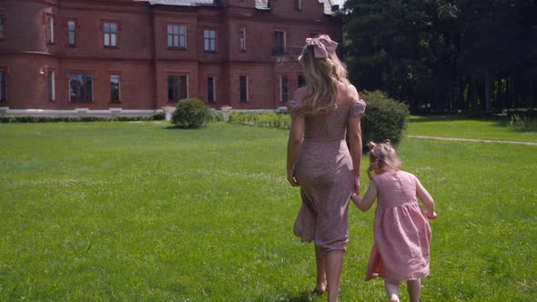 Woman and a Child in Pink Dresses Run Across a Green Field Against the Background of an Old Castle