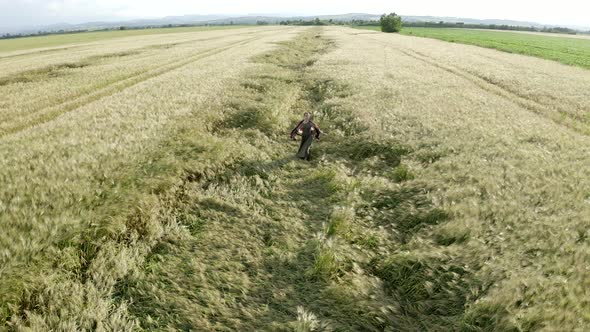 Aerial view young lady in long dress, shawl walking in agricultural wheat field at sunset, harvest