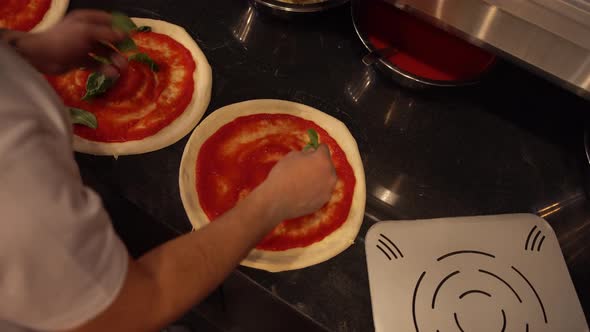 Professional Cook Adding Basil Leaves on the Pizza Dough Covered in Tomato Sauce in Restaurant
