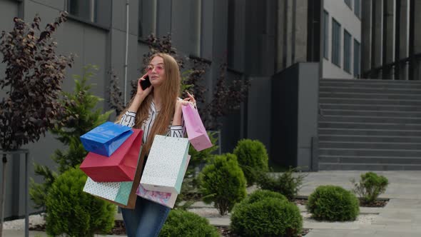 Positive Young Girl with Bags Talking on Mobile Phone About Sales in Shopping Mall in Black Friday