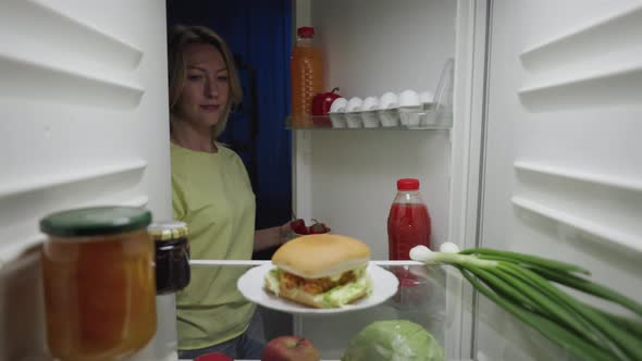 Young Woman Opens the Refrigerator Door at Night and Happy Takes a Burger
