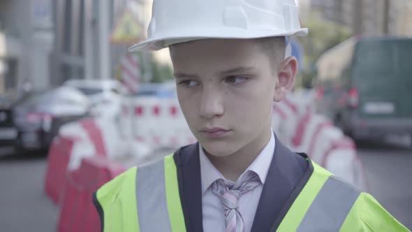 Portrait of Sad Tired Little Boy in Constructor Helmet on His Head, and Uniform Looking Away