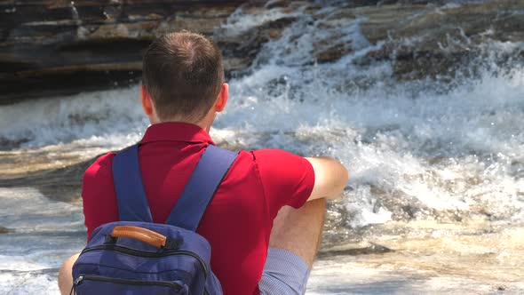 Back View of a Male Traveler in a Red Polo Sitting By a Mountain River