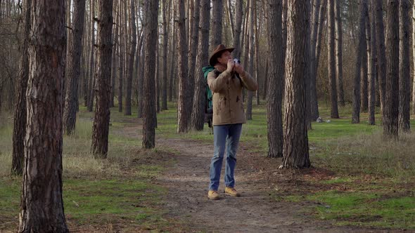 A Man in a Hat with a Backpack Takes Pictures in the Forest