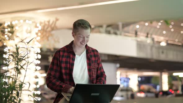 Young Hipster Man Thinking And Smiling While Using Laptop In The City At Night