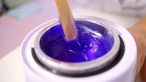 A Female Hand in Red Gloves Stirs the Melted Blue Wax with a Wooden Stick for Depilation