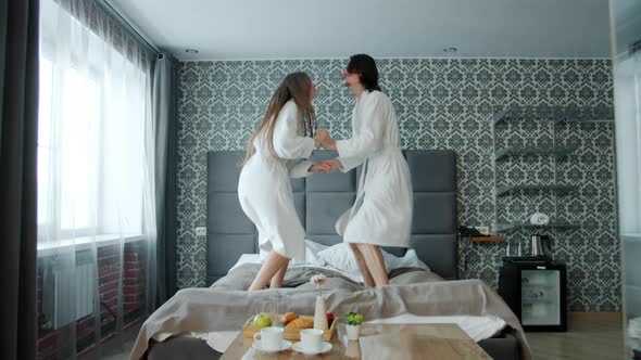 Happy Husband and Wife Wearing Bathrobes Jumping on Bed in Hotel Room Having Fun